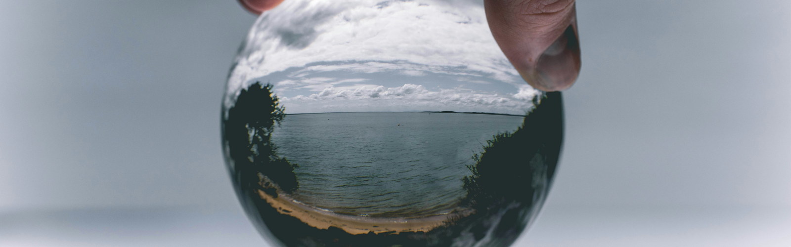 A hand holding a glass lens ball, with the coastline behind it appearing inverted inside the glass.
